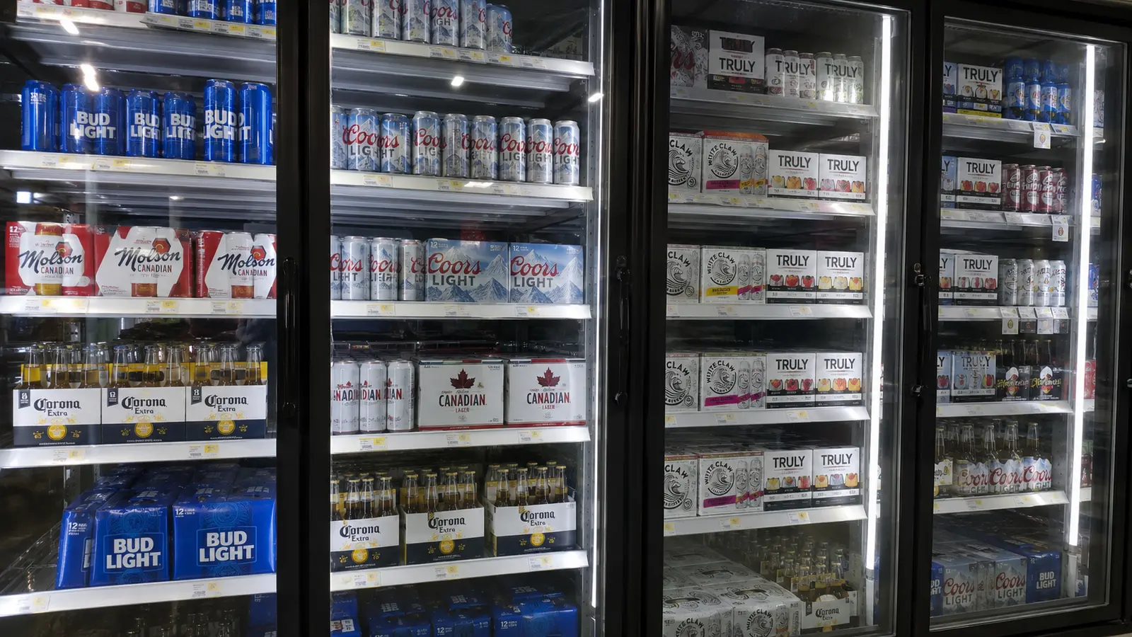 A convenience store beer cooler shot through the glass doors — stacked cases of Bud Light, Coors Light, Molson Canadian and Corona Extra alongside White Claw and Truly seltzers, every one of them a familiar low-ABV brand and nothing on the shelf above the 7.1% limit